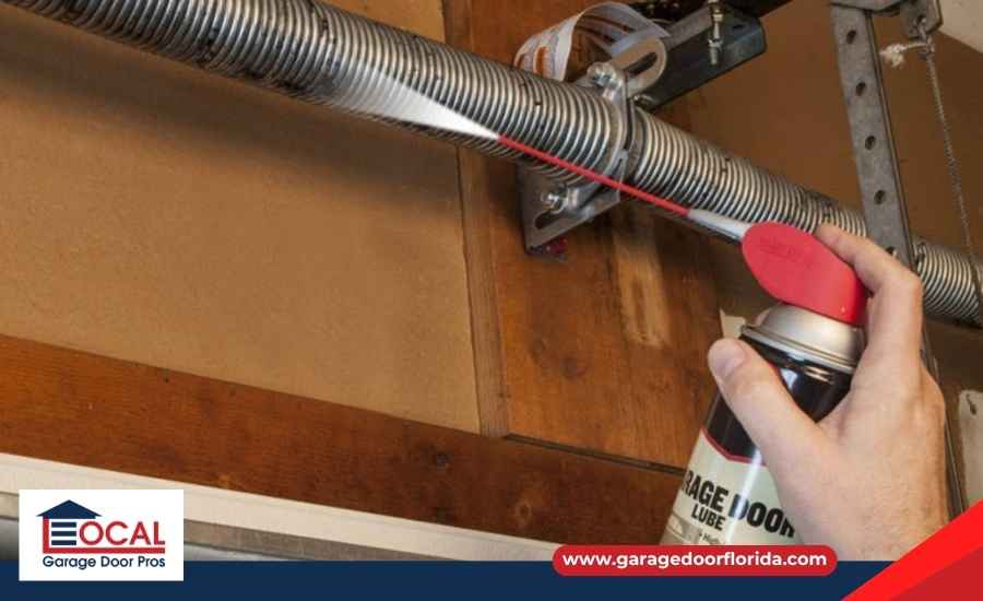 A homeowner applying spray lubricant to the metal hinges and track mechanisms of an open garage door.