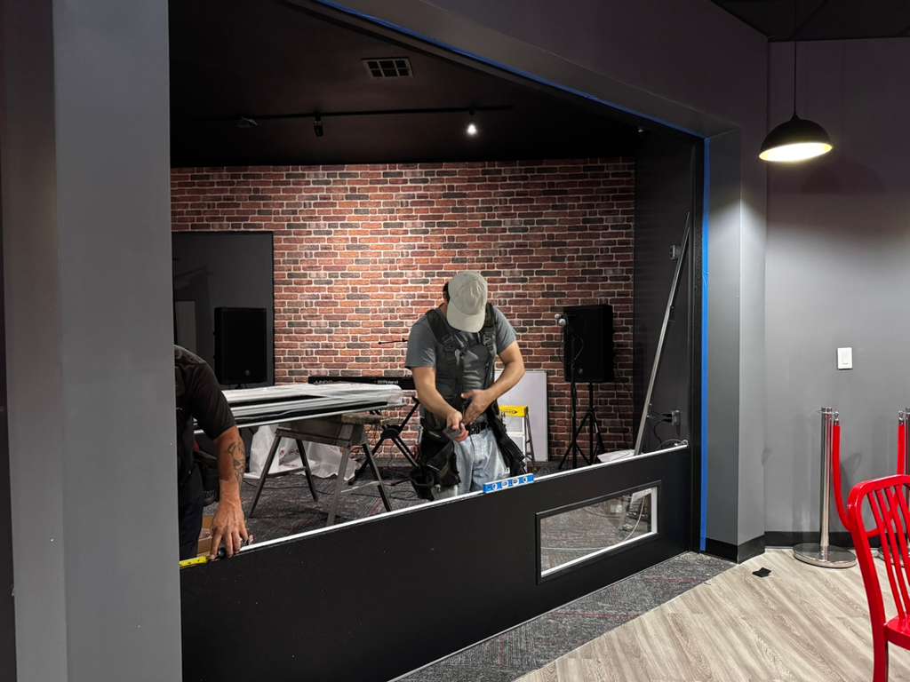Worker in a gray shirt and cap stands in a brick-walled studio area, adjusting tools near a black counter and equipment.
