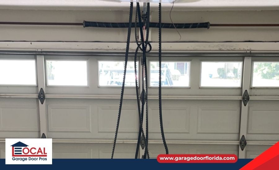 A broken black drive belt hanging down from a garage door opener mechanism in front of a closed white garage door.